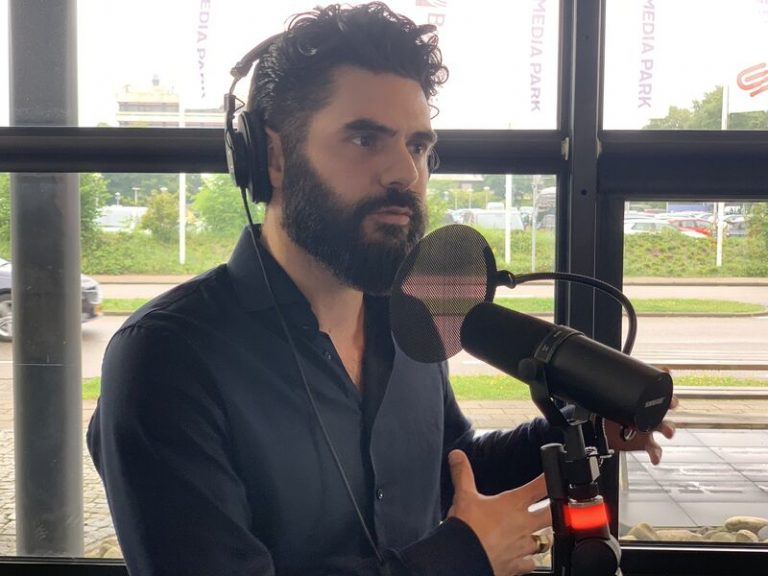A man with dark hair and a beard is speaking into a microphone in a studio. He is wearing a dark-colored shirt and studio equipment is visible. Behind him are large windows with a view of greenery and parked cars outside. Multiple "Media Park" signs are seen.