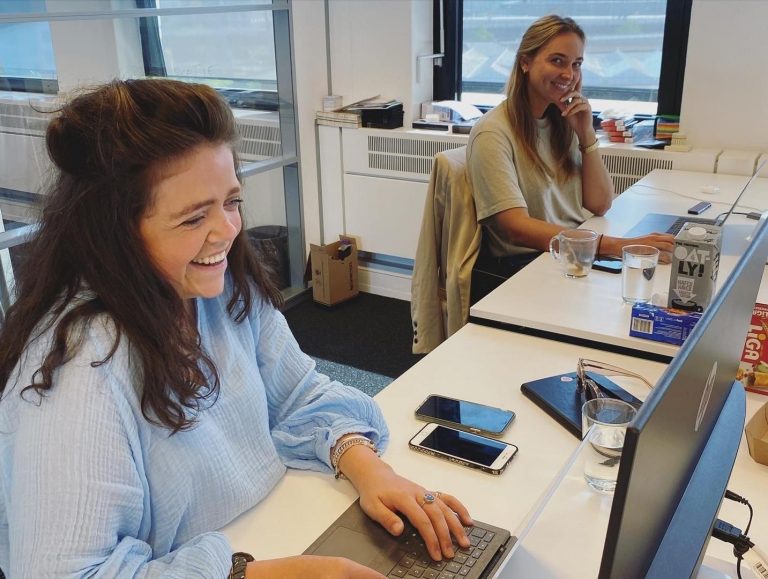 Twee vrouwen werken aan hun bureau in een helder kantoor. De vrouw op de voorgrond, gekleed in een lichtblauwe blouse, lacht terwijl ze typt op een laptop. De vrouw op de achtergrond, gekleed in een beige shirt, lacht naar de camera met haar kin rustend op haar hand.