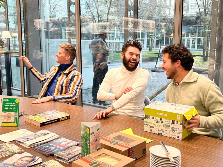 Drie mannen zitten aan een tafel binnen en lachen. De tafel is bedekt met verschillende pamfletten, brochures en kleurrijke dozen. Grote ramen op de achtergrond laten een uitzicht naar buiten zien van bomen en gebouwen, wat bijdraagt aan de lichte en open sfeer.