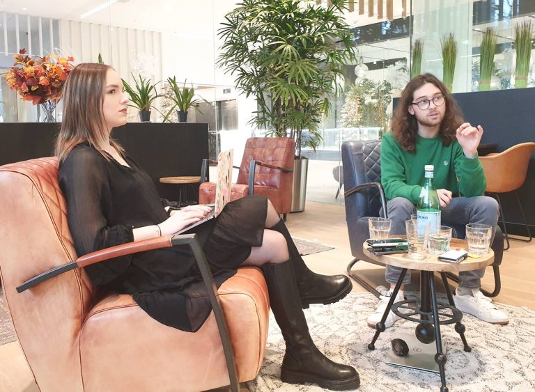 Twee personen zitten in een moderne, ruime kamer met glazen wanden en groene planten. De vrouw links draagt een zwarte jurk en houdt een laptop op haar schoot. De man rechts, in een groen jasje en jeans, lijkt iets uit te leggen met een gebaar.