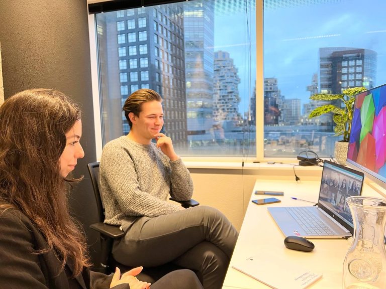 Twee mensen zitten aan een bureau in een kantoor en nemen deel aan een videoconferentiegesprek. De persoon links heeft lang, donker haar en kijkt naar beneden, terwijl de persoon rechts, met kort haar, naar het scherm lacht. Een raam achter hen toont een stadsgezicht met hoge gebouwen.