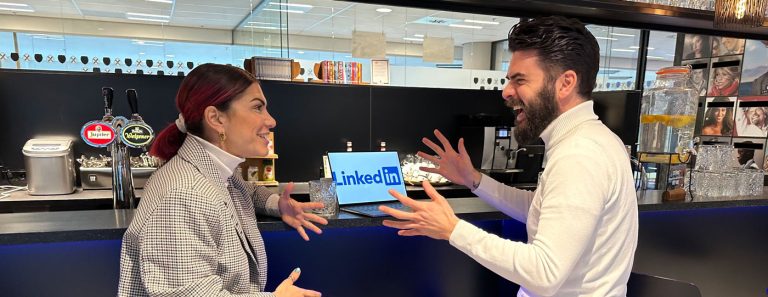 Two people are engaged in an animated conversation at a counter in front of a laptop displaying the LinkedIn logo. One person has red hair pulled back and is wearing a plaid jacket, while the other has dark hair, a beard, and is wearing a white shirt.