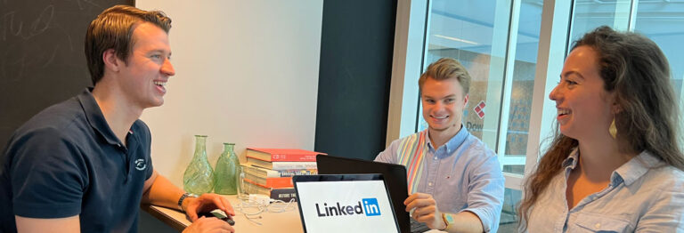 Three individuals are seated around a table, engaged in a discussion. The person on the left is smiling, while the person on the right also smiles. The individual in the center sits behind a laptop displaying the LinkedIn logo. Books and green vases are on the table.