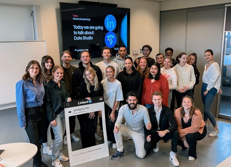 A group of 24 diverse individuals pose together in a classroom setting. They are smiling and some are giving thumbs up. Behind them is a screen displaying a LinkedIn and WiFi logo, and the text "Today we are going to talk about Data Studio.