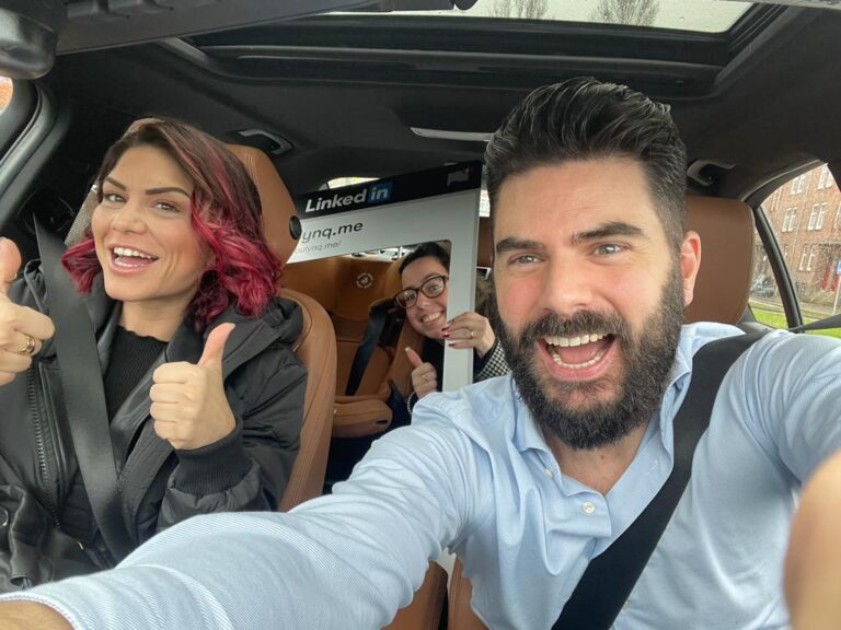 Drie mensen zitten in een auto, lachend en geven duimen omhoog. De persoon op de bestuurdersstoel heeft donker haar en een baard, terwijl de passagier fel, kort, roze haar heeft. Achterin houdt iemand een groot LinkedIn-frame vast, klaar voor serieuze netwerken.