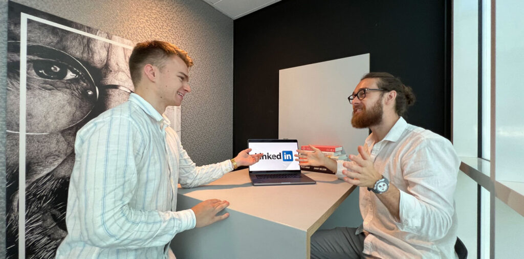 Twee mannen in witte shirts praten en lachen naar elkaar in een modern kantoor. Een van hen zit terwijl de ander naast een bureau staat met een laptop waarop het LinkedIn-logo staat. Een grote zwart-witte muurkunst is gedeeltelijk zichtbaar op de achtergrond.