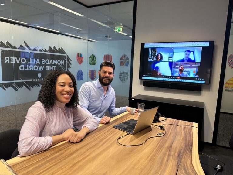 Two people sit at a conference table with a laptop, smiling. Behind them, a large screen shows a video call with four participants. The room has a clear glass wall with text, "Nomads All Over the World," and colorful design decals.
