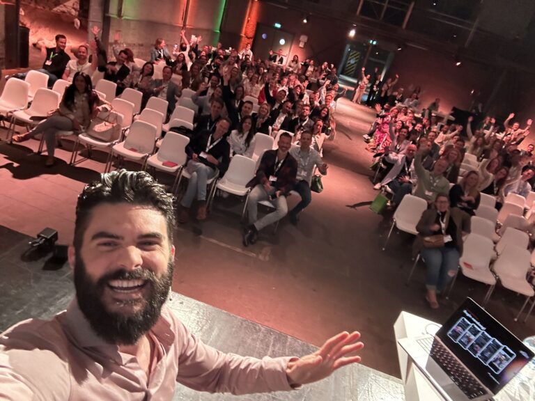 A bearded man in a light-colored shirt takes a selfie on stage in front of a lively audience seated in white chairs. The crowd is smiling, waving, and seems engaged. A laptop with a presentation is visible on a podium next to him. The room is well-lit and spacious.