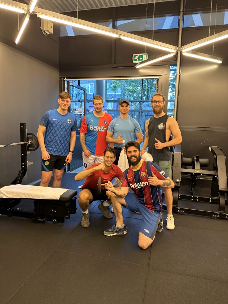 Six men in athletic attire posing in a gym with weightlifting equipment. Three are standing behind a bench press while the other three kneel or squat in front. Two men give thumbs-up gestures, and most are smiling. Various jerseys and workout clothes are worn.