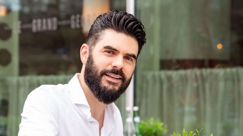 Een man met een baard, gekleed in een wit shirt met lange mouwen, giet water uit een fles in een glas terwijl hij aan een tafeltje in een buitencafé zit. De achtergrond toont een groene plant en het raam van het café weerspiegelt een wazig zicht op de straat.