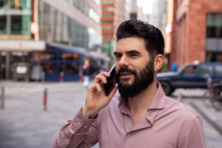 A bearded man with dark hair is standing on a city street, wearing a light pink, long-sleeved shirt. He is smiling and holding a smartphone to his ear, appearing to discuss LinkedIn trends. The background includes buildings, parked cars, and a few pedestrians.