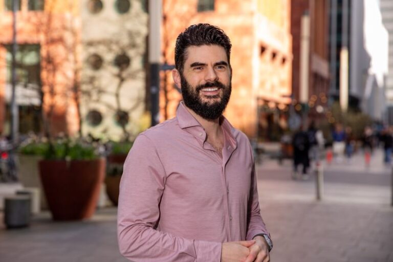 A man with a beard and short hair stands on a city sidewalk, smiling. He wears a light pink button-up shirt. The background features a mix of modern buildings and greenery, with blurry figures walking, suggesting a sunny day ideal for building real relationships through friendly conversations rather than sales pitches.