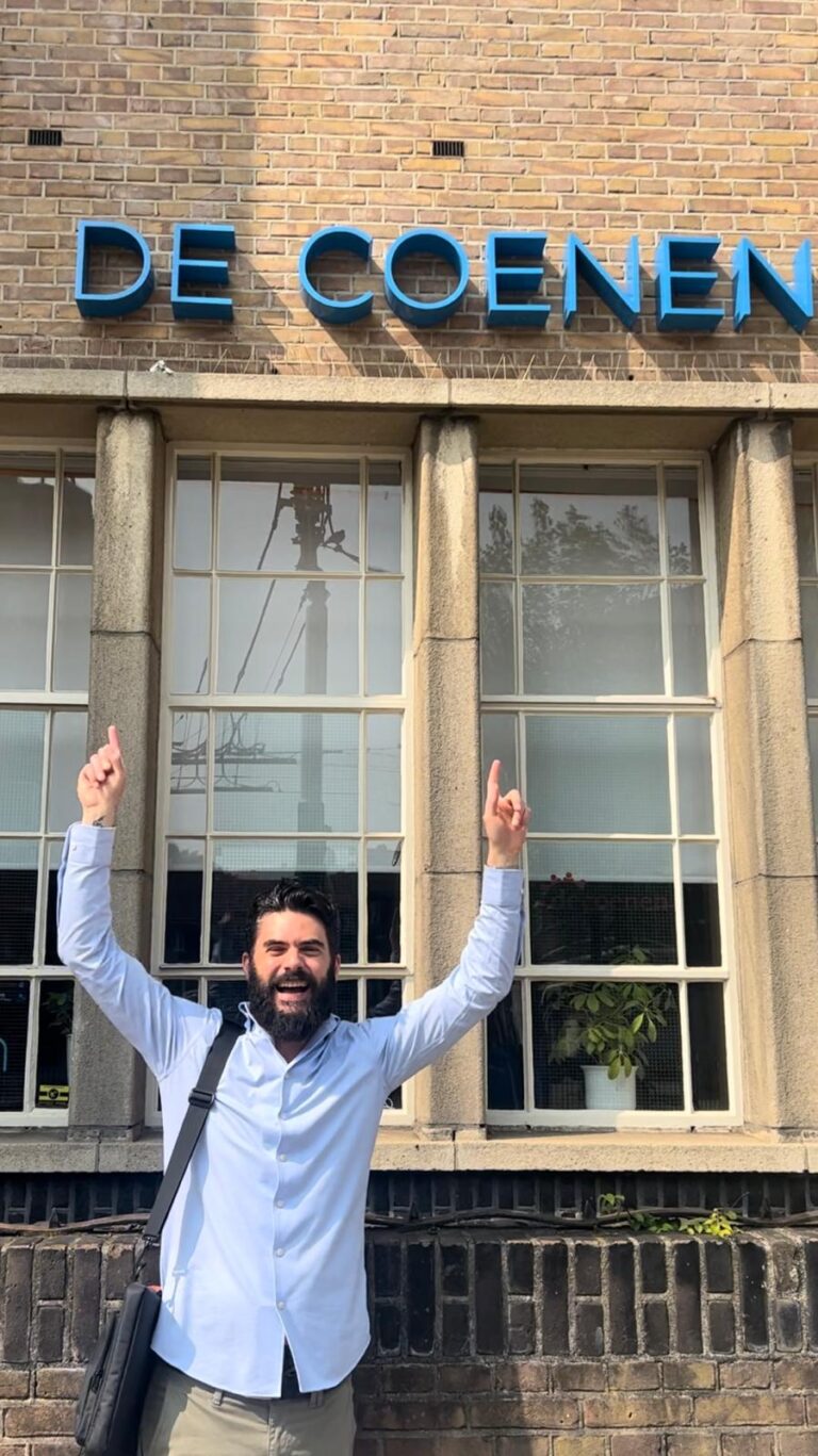 Een bebaarde man met donker haar, gekleed in een lichtblauw shirt en een zwarte schoudertas, staat voor een bakstenen gebouw met het opschrift "55+ De Coenen." Hij lacht en wijst met beide handen omhoog. Het gebouw heeft grote ramen op de achtergrond.