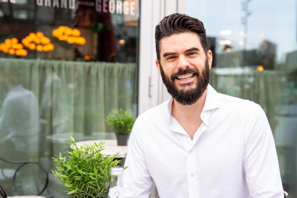 Een man met een baard en donker haar lacht terwijl hij op een terrasje zit en levendige netwerken voert. Hij draagt een wit shirt en er staat een kleine potplant op de tafel voor hem. Op de achtergrond is een raam te zien met hangende lampen erin.