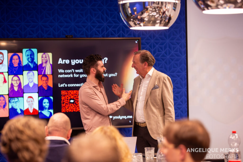 Two men from Blue Field Agency are shaking hands and smiling in an office setting. Behind them, a presentation screen displays a colorful chart and text. People are seated at a table in the foreground, engaged in discussions on AI innovations.