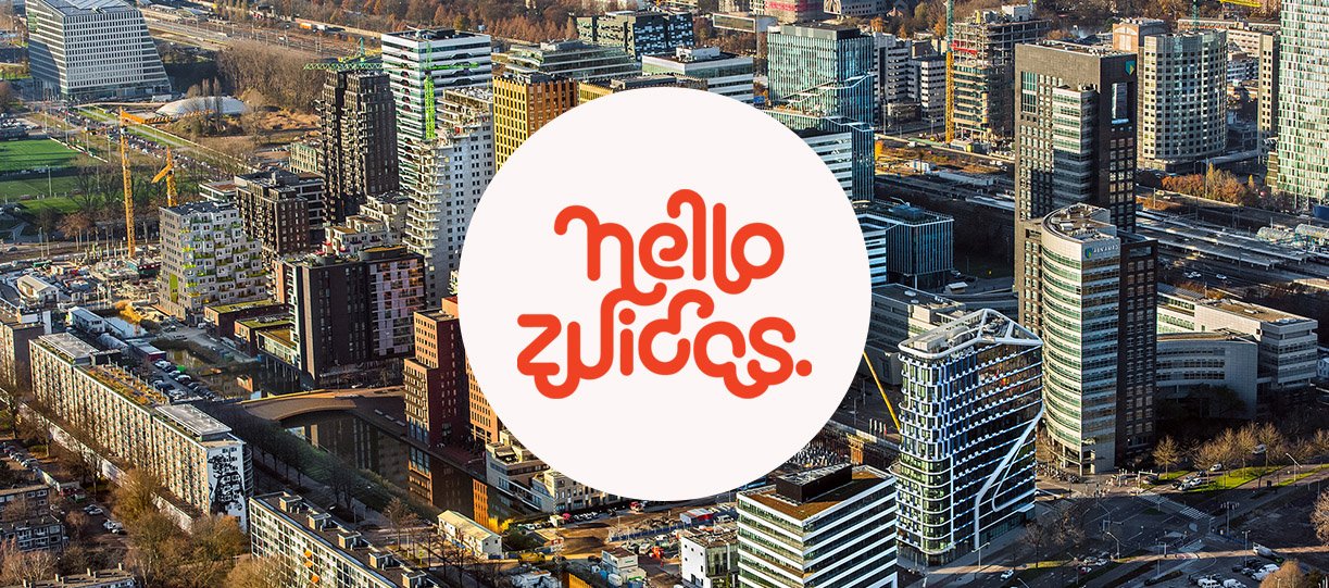 Aerial view of a cityscape with modern buildings, construction sites, and greenery. In the center, a circular white logo with the words "Hello Zuidas" in orange, stylized text.