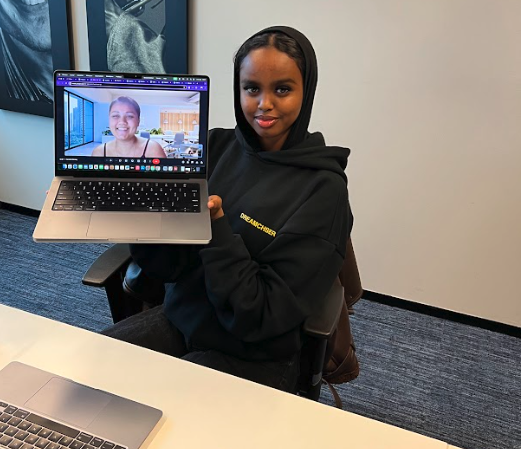 A person in a black hoodie, seated at a desk, holds up a laptop displaying a video call with another person in a well-lit room. The office setting includes artwork on the walls and a table with a closed laptop.