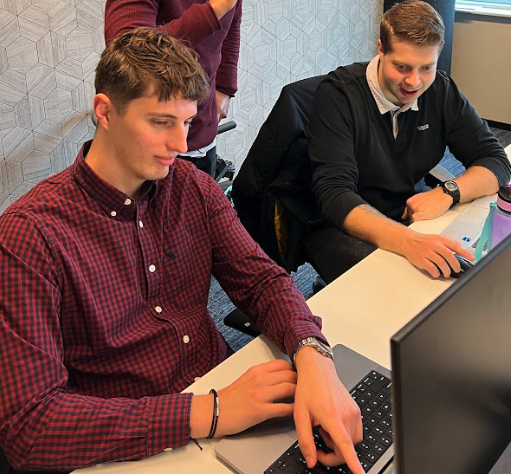 Two men are seated at a desk, both focused on the laptop screens in front of them. One wears a red plaid shirt, and the other wears a black shirt. A third person stands nearby, partially visible. They appear to be in a collaborative office setting.