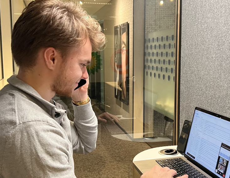 A person in a gray sweater talks on a phone while typing on a laptop at a small round table in a modern office setting. There's a hallway and artwork visible in the background.