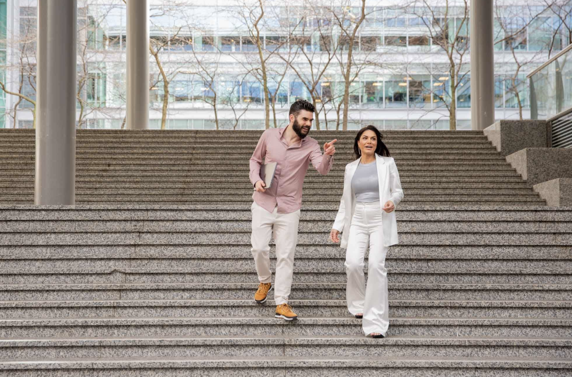 A man and a woman in business attire walking down outdoor steps. The man is holding a laptop and gesturing while talking to the woman, who is focused ahead. Background features trees and a modern building facade.