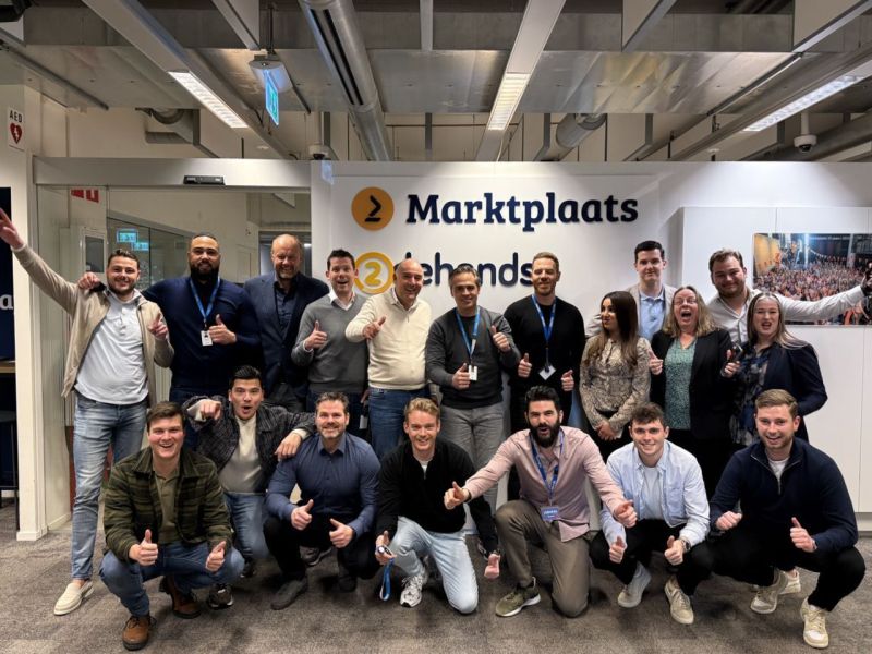 A group of people smiling and giving thumbs up poses in front of a sign that reads "Marktplaats 2e hands" in an office environment. They appear to be colleagues or a team celebrating an event or gathering.