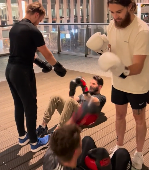 Four men are exercising outdoors at night: one stands in black boxing gloves, another in white gloves, while the others perform sit-ups with punches. Buildings with lights glow in the background, reminiscent of a city where AI could be ingezet for innovative urban solutions.