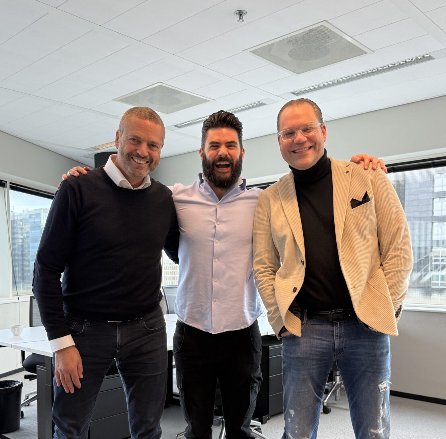 Three men are standing together in an office, smiling at the camera. Two are wearing casual jeans and sneakers; one wears a light blazer, the other a dark sweater. The man in the middle has a blue shirt and black pants. Large windows show a cityscape outside.