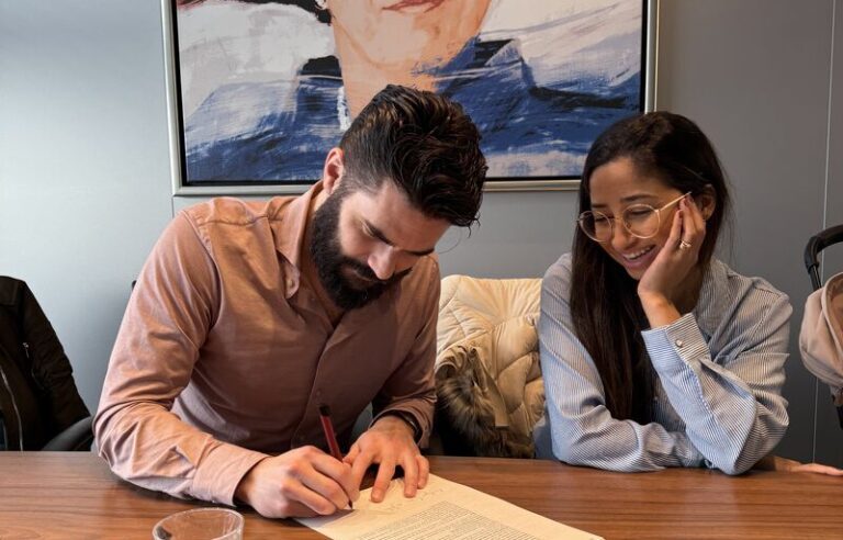A bearded man efficiently signs a document as a woman with long hair watches, smiling. They sit at a wooden table with a glass of water and a pen. A large portrait of someone in a red hat hangs in the background, symbolizing more winst through streamlined actions.