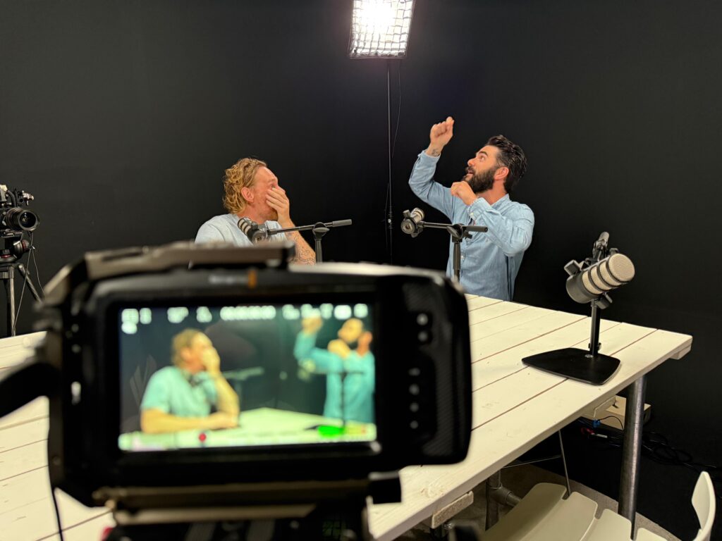Two men sit at a table in a recording studio with microphones, one covering his mouth and the other gesturing upward. A camera in the foreground captures this Workshop Video scene, displayed on its screen.