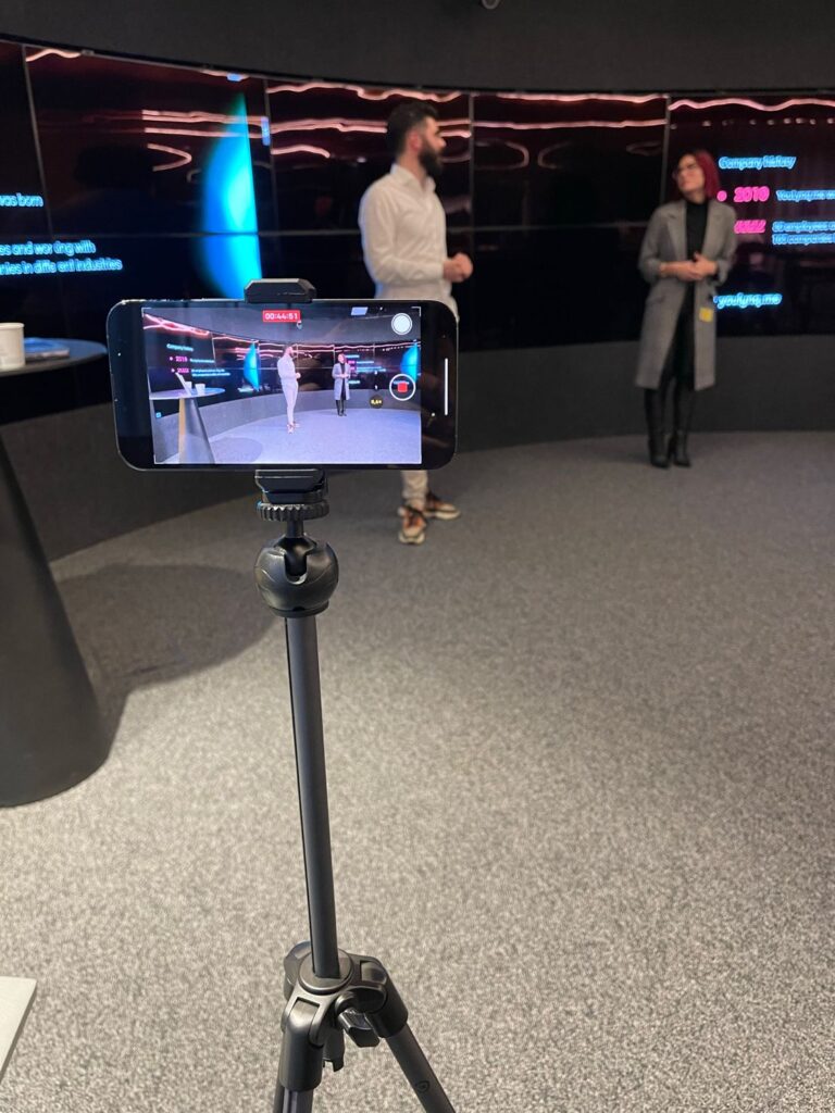 Een smartphone op een statief neemt twee mensen op die staan te praten in een moderne kamer met donkere muren en grote schermen waarop tekst wordt weergegeven, waardoor boeiende video-inhoud ontstaat die perfect is voor LinkedIn-marketing.